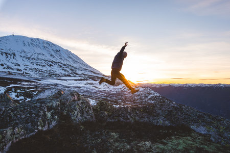 Traveler jumps from rocks at sunset near Gaustatoppen Mountain in Norway. Golden light, snow peaks, and open sky capture pure joy and freedom of adventure.の写真素材