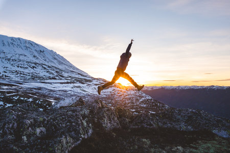 Traveler jumps from rocks at sunset near Gaustatoppen Mountain in Norway. Golden light, snow peaks, and open sky capture pure joy and freedom of adventure.の写真素材