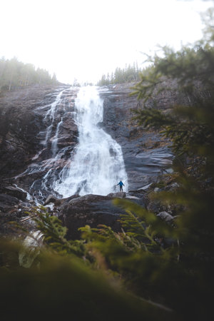 Person standing below powerful Reiarsfossen waterfall in Setesdal Valley, Norway. Dramatic scene showing human scale against raw Nordic nature and water energy.の写真素材