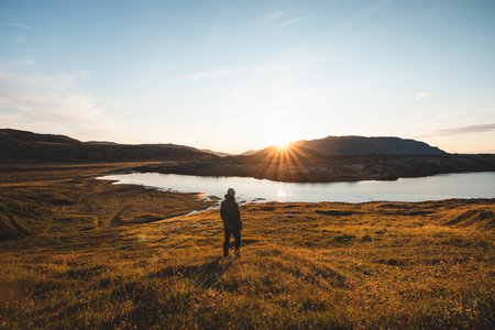 Lone hiker watches the sun set over a peaceful mountain lake, surrounded by golden grass and rugged Icelandic wilderness