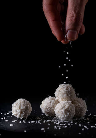 sprinkling coconut on some coconut bonbons, on a black background. cake shop.