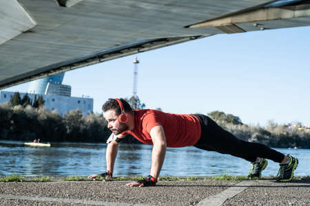young man doing push-ups with a red t-shirt and music headphones, in an urban environment. healthy living conceptの写真素材