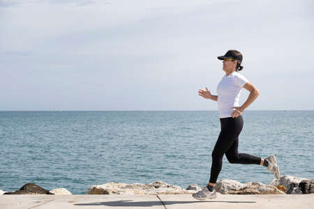 woman with cap and sunglasses running along the promenade, the sea in the background