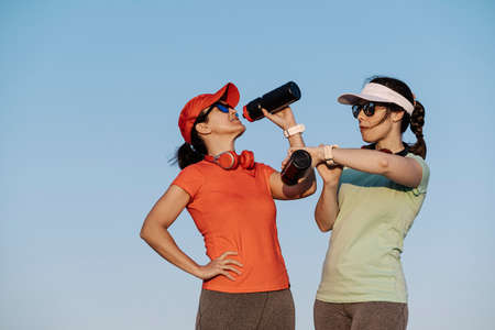 two women hydrating themselves with water and looking at their watch after runningの写真素材