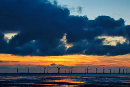 Sunset over sea with silhouette of offshore wind turbines, vibrant sky, and reflection on water in...
