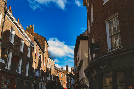 Charming European street scene with historic brick buildings under a clear blue sky with fluffy clouds, showcasing architectural details and local businesses in York, North Yorkshire, England.の写真素材