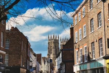 Historic European city street with traditional brick buildings and a prominent Gothic cathedral in the background under a blue sky with clouds in York, North Yorkshire, England.の写真素材