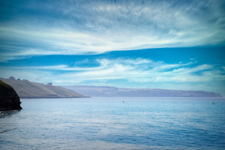 A serene coastal scene with calm blue waters, a distant shoreline, and a partly cloudy sky. The...