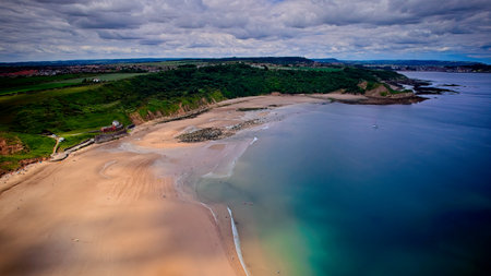 Aerial view of a serene beach with golden sand, clear blue water, and lush green hills in the background under a cloudy sky.の写真素材