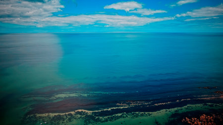 Aerial view of a serene ocean with clear blue water and a partly cloudy sky. The shoreline is...
