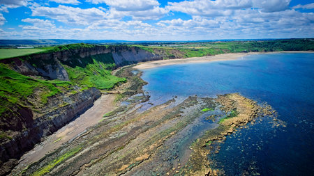 Aerial view of a coastal landscape with cliffs, a sandy beach, and clear blue waters under a partly cloudy sky.の写真素材