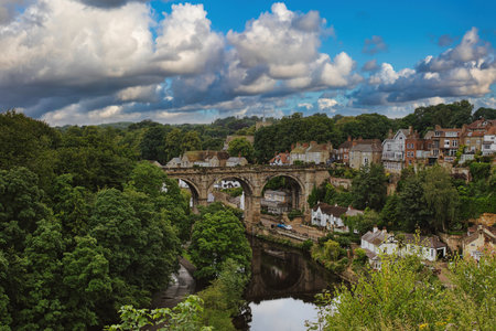 A scenic view of a historic stone bridge over a river, surrounded by lush greenery and quaint houses...
