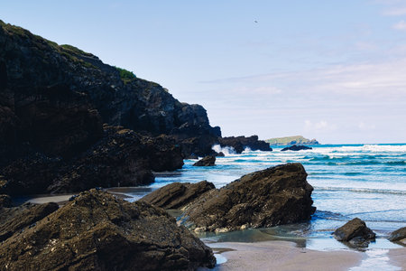 A scenic coastal view featuring rocky cliffs and a sandy beach. Waves gently crash against the dark rocks, with a clear blue sky and distant hills in the background.の写真素材