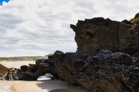 A rocky beach landscape featuring large, dark rocks with a textured surface. The sky is partly cloudy, creating a dramatic backdrop. The sandy beach is visible in the foreground, with a hint of distant hills and a small town in the background.の写真素材