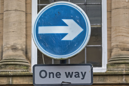 A circular blue road sign with a white right arrow, indicating one-way traffic. Below it, a rectangular sign reads 'One way'. The background features a historic building with stone columns.の写真素材