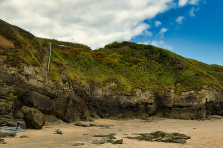 A scenic view of a rocky beach with a steep grassy cliff. The cliff is adorned with greenery and has...