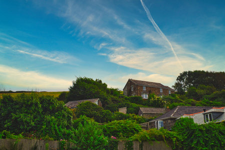 A picturesque scene of a countryside house surrounded by lush greenery and trees under a blue sky with wispy clouds. The house is nestled on a hillside, showcasing a tranquil rural setting.の写真素材