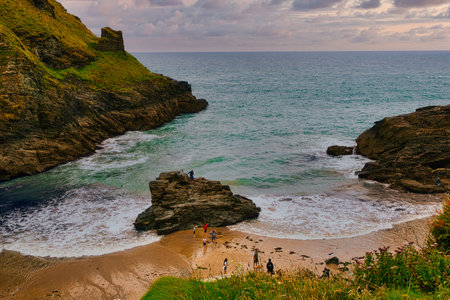 A scenic coastal view featuring a sandy beach surrounded by rocky cliffs. People are enjoying the beach, with gentle waves lapping at the shore. The sky is partly cloudy, adding to the serene atmosphere.の写真素材