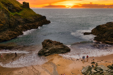 A scenic beach at sunset, featuring rocky cliffs and gentle waves. People stroll along the shore, enjoying the tranquil atmosphere. The sky is painted in warm hues of orange and yellow as the sun sets over the horizon.の写真素材