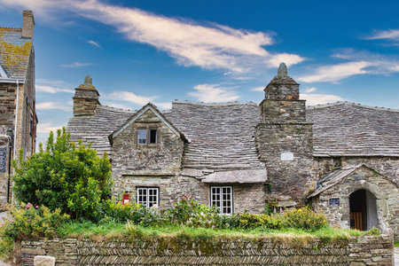 A charming stone cottage with a slate roof, surrounded by lush greenery and a well-maintained garden. The sky is bright with scattered clouds, adding to the picturesque scene.の写真素材