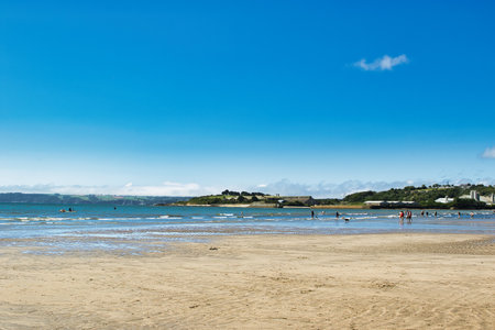 A serene beach scene with golden sand, gentle waves, and a clear blue sky. People are enjoying various activities in the water, while green hills and buildings are visible in the background.の写真素材