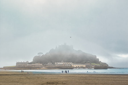 A misty view of a coastal landscape featuring a castle on a hill, surrounded by fog. The foreground shows a beach with people walking, while the background reveals the castle partially obscured by clouds.の写真素材