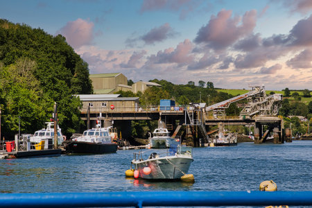 A scenic view of a harbor with boats moored in the foreground. The background features a dock with industrial buildings and lush greenery. The sky is partly cloudy, creating a picturesque atmosphere.の写真素材
