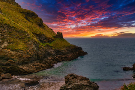 A stunning coastal landscape at sunset, featuring rocky cliffs and a vibrant sky filled with orange, pink, and purple hues. The calm sea reflects the colors of the sky, while a small figure stands on the beach.の写真素材