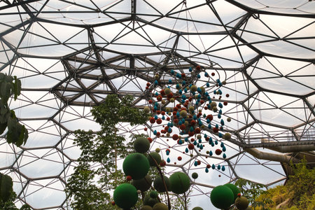 A view of a geodesic dome structure with a colorful hanging sculpture made of spheres. The dome is filled with greenery, showcasing a blend of nature and modern architecture.の写真素材