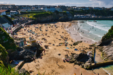 A vibrant beach scene with numerous people enjoying the sun and sand. The beach is dotted with colorful umbrellas and towels, while waves gently lap at the shore. In the background, there are houses and cliffs, creating a picturesque coastal view.の写真素材