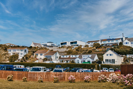 A scenic view of a hillside residential area with various houses and parked cars. The foreground features a wooden fence and colorful flowers, while the background showcases a clear blue sky and green hills.の写真素材