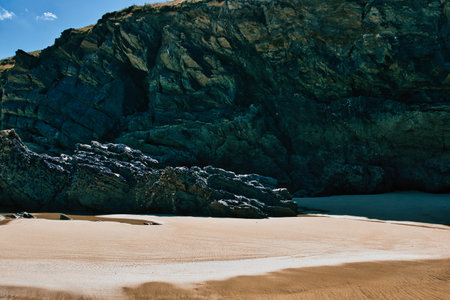 A serene beach scene featuring smooth golden sand and rugged dark rocks. The backdrop consists of a steep rocky cliff with a mix of textures and colors, under a clear blue sky with a few clouds. The tranquil atmosphere invites relaxation and exploration.の写真素材