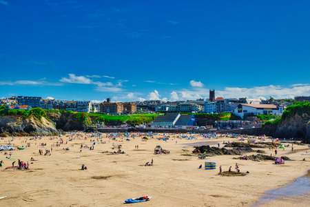 A vibrant beach scene with numerous people enjoying the sun and sand. The beach is expansive, with colorful umbrellas and towels scattered across the golden sand. In the background, there are buildings and a clear blue sky with a few clouds.の写真素材