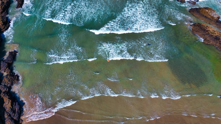 Aerial view of a beach with waves crashing onto the shore. The water is a mix of turquoise and...