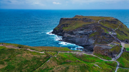 Aerial view of a rugged coastal landscape featuring a rocky cliff with lush greenery and pathways leading to the edge. The ocean is visible in the background, with waves crashing against the rocks. The sky is partly cloudy, creating a serene atmosphere.の写真素材