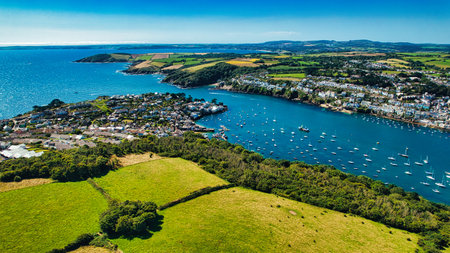 Aerial view of a coastal town surrounded by lush green fields and a serene blue bay filled with boats. The landscape features rolling hills and a clear sky, showcasing the beauty of nature and the charm of the seaside community.の写真素材