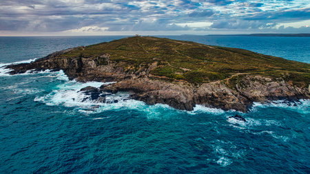 Aerial view of a rocky island surrounded by turquoise waters, with waves crashing against the shore. The island features green vegetation and a path leading to a distant lighthouse on the top. The sky is partly cloudy, creating a dramatic atmosphere.の写真素材