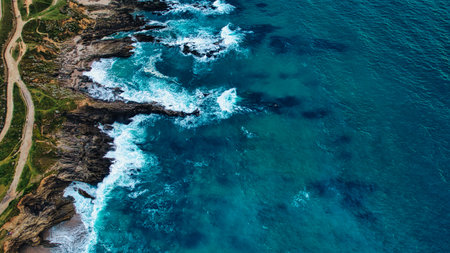 Aerial view of a rocky coastline with crashing waves and clear blue water. The shoreline features...