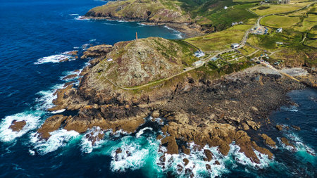 Aerial view of a rugged coastline featuring rocky cliffs, crashing waves, and a lighthouse on a hill. The landscape includes green fields and a small village, with the ocean in vibrant shades of blue and turquoise.の写真素材