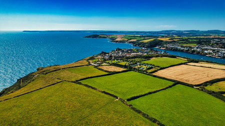 Aerial view of a coastal landscape featuring lush green fields, a small town by the water, and a serene blue sea. The scene captures the beauty of nature with rolling hills and agricultural land.の写真素材