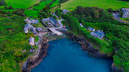 Aerial view of a coastal village with lush green hills, rocky shoreline, and a small harbor. The scene includes several buildings, a parking area, and boats in the water, surrounded by vibrant greenery.の写真素材
