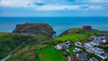 Aerial view of a coastal landscape featuring a rocky outcrop, lush green hills, and a small village by the sea. The ocean is visible in the background, with a cloudy sky overhead.の写真素材