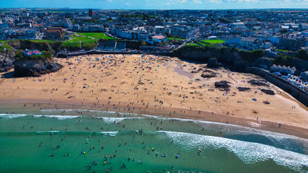 Aerial view of a busy beach with golden sand and turquoise water. Numerous people sunbathing, swimming, and surfing. In the background, a coastal town with buildings and green fields. Clear blue sky with a few clouds.の写真素材