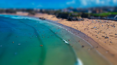 Aerial view of a beach with many people enjoying the sun and water. The scene features a sandy shore, waves crashing, and surfers in the water. The background includes green hills and buildings, creating a vibrant coastal atmosphere.の写真素材