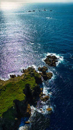 Aerial view of a rocky coastline with lush green vegetation, surrounded by deep blue ocean waters. The sunlight reflects off the water, creating a shimmering effect. In the distance, a lighthouse can be seen on a small island.の写真素材
