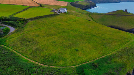 Aerial view of a green landscape with a solitary house near the coast. The area features rolling hills, agricultural fields, and a winding road leading to the house. The ocean is visible in the background, creating a serene coastal scene.の写真素材