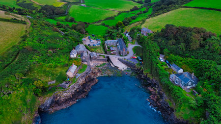 Aerial view of a coastal village surrounded by lush green fields and rocky shores. The village features quaint buildings, a small harbor, and a winding road leading to the water. The landscape is vibrant with greenery and the ocean is calm.の写真素材