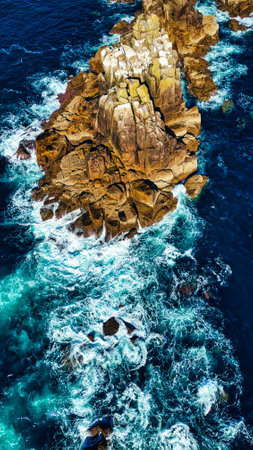 Aerial view of rugged coastal rocks surrounded by turquoise ocean waves. The water crashes against the rocky formations, creating white foam. The scene captures the beauty of nature with a vibrant contrast between the rocks and the sea.の写真素材