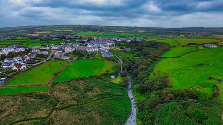 Aerial view of a picturesque village surrounded by lush green fields and hills under a cloudy sky. The village features white and colorful houses, with a winding road leading through the landscape. The scene captures the beauty of rural life in a serene environment.の写真素材