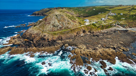 Aerial view of a rugged coastline featuring rocky cliffs, crashing waves, and a lighthouse on a hill. The landscape is lush and green, with a winding road and scattered white houses in the background. The ocean is a vibrant blue, contrasting with the rocky shore.の写真素材